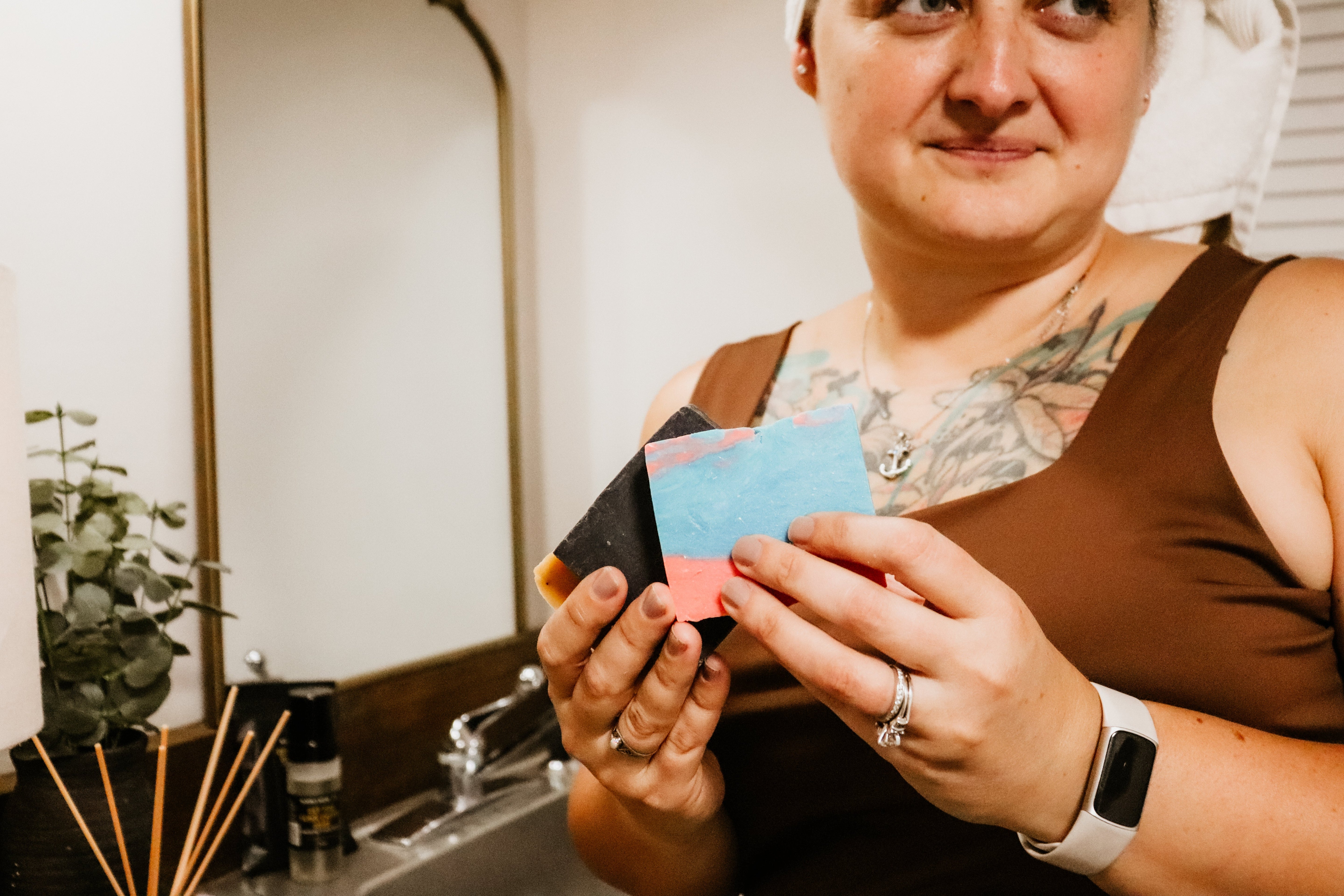 Rebecca Mitchell holding natural soap bars in various colors, featuring visible ingredients, in a bathroom setting with a towel wrapped around her head—spa-like, self-care moment.