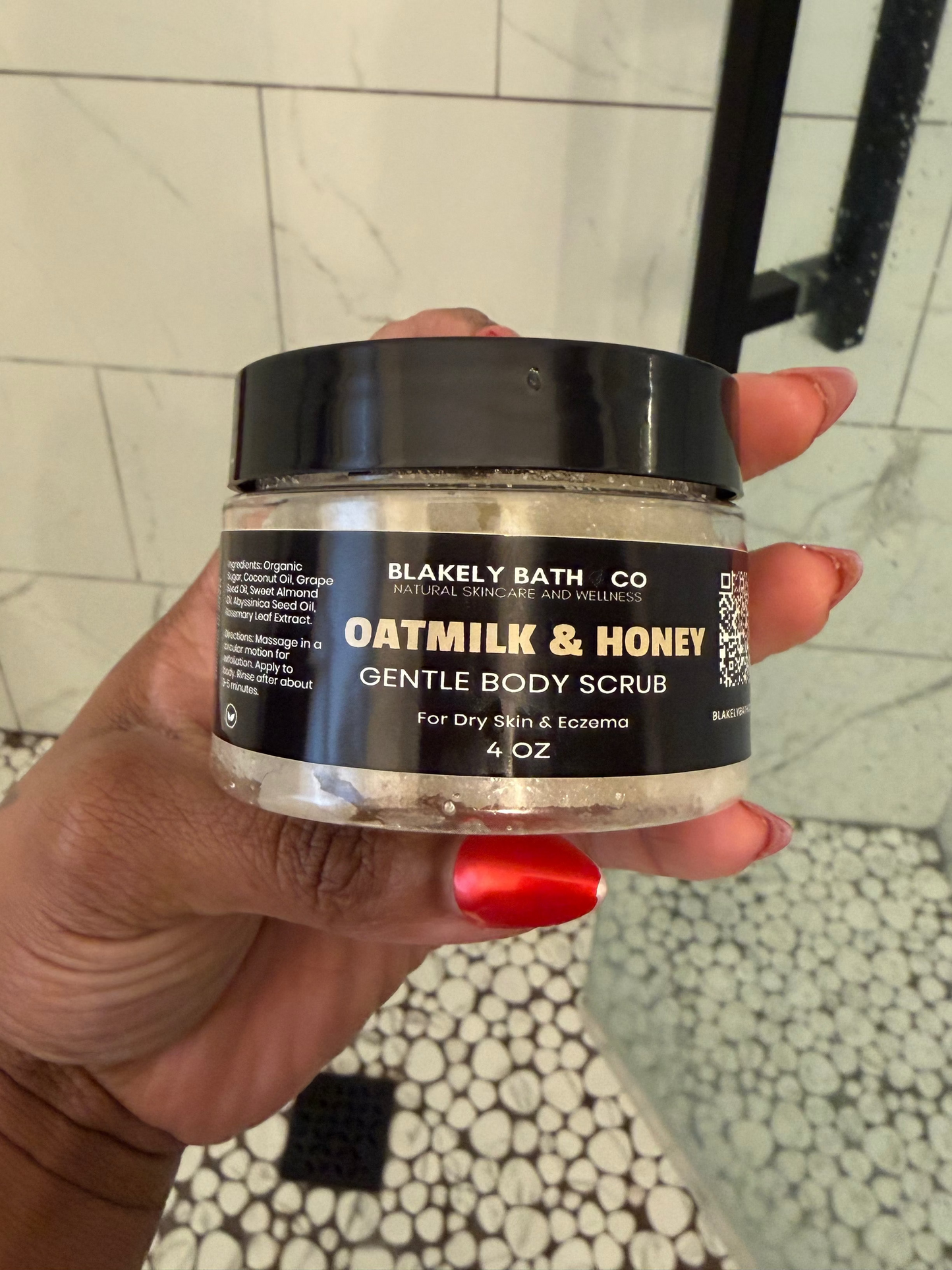 Hand holding a jar of Oatmilk & Honey Gentle Body Scrub from Blackely Bath Co in a bathroom setting.