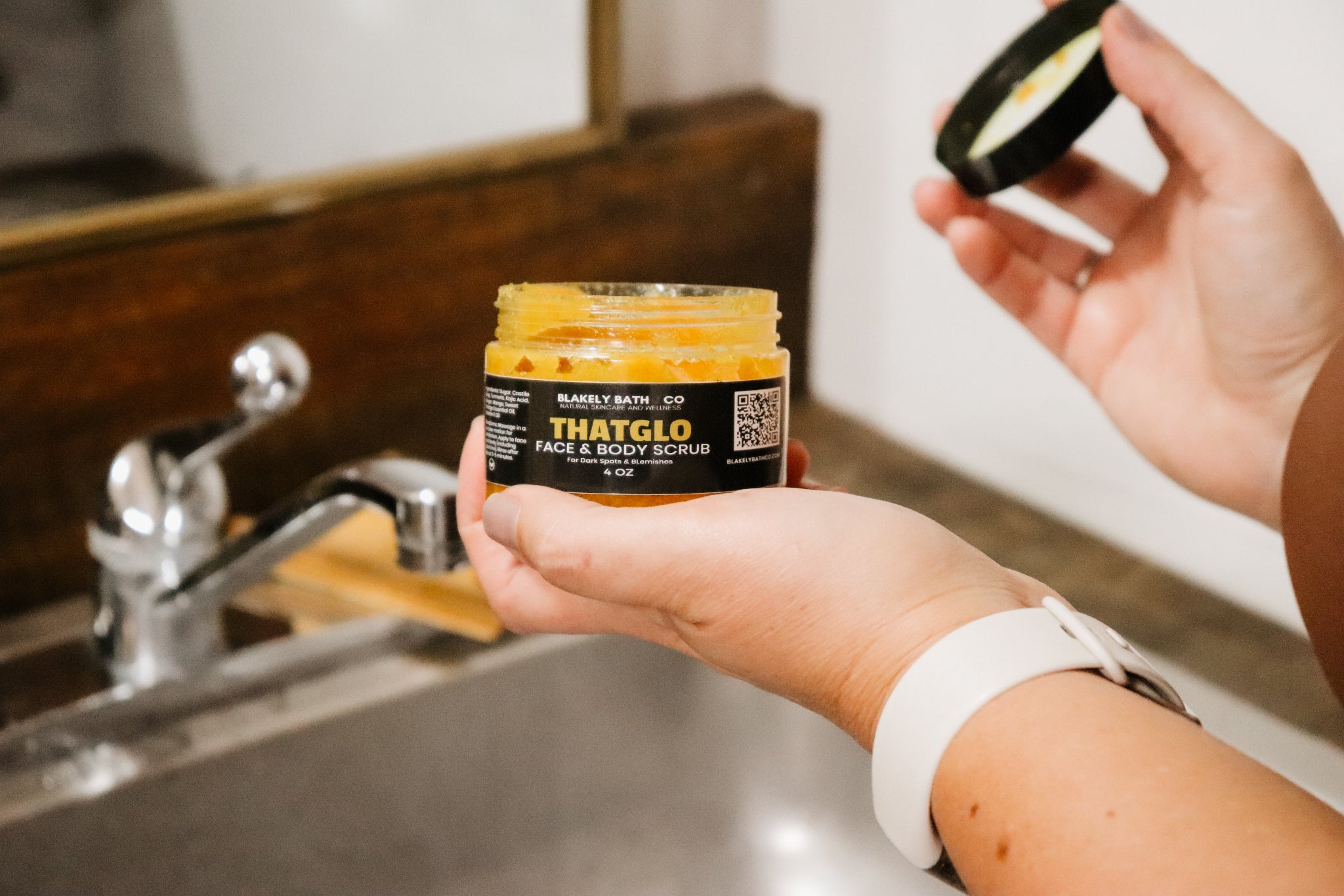 Woman holding a jar of THATGLO turmeric face and body scrub in a bathroom setting.