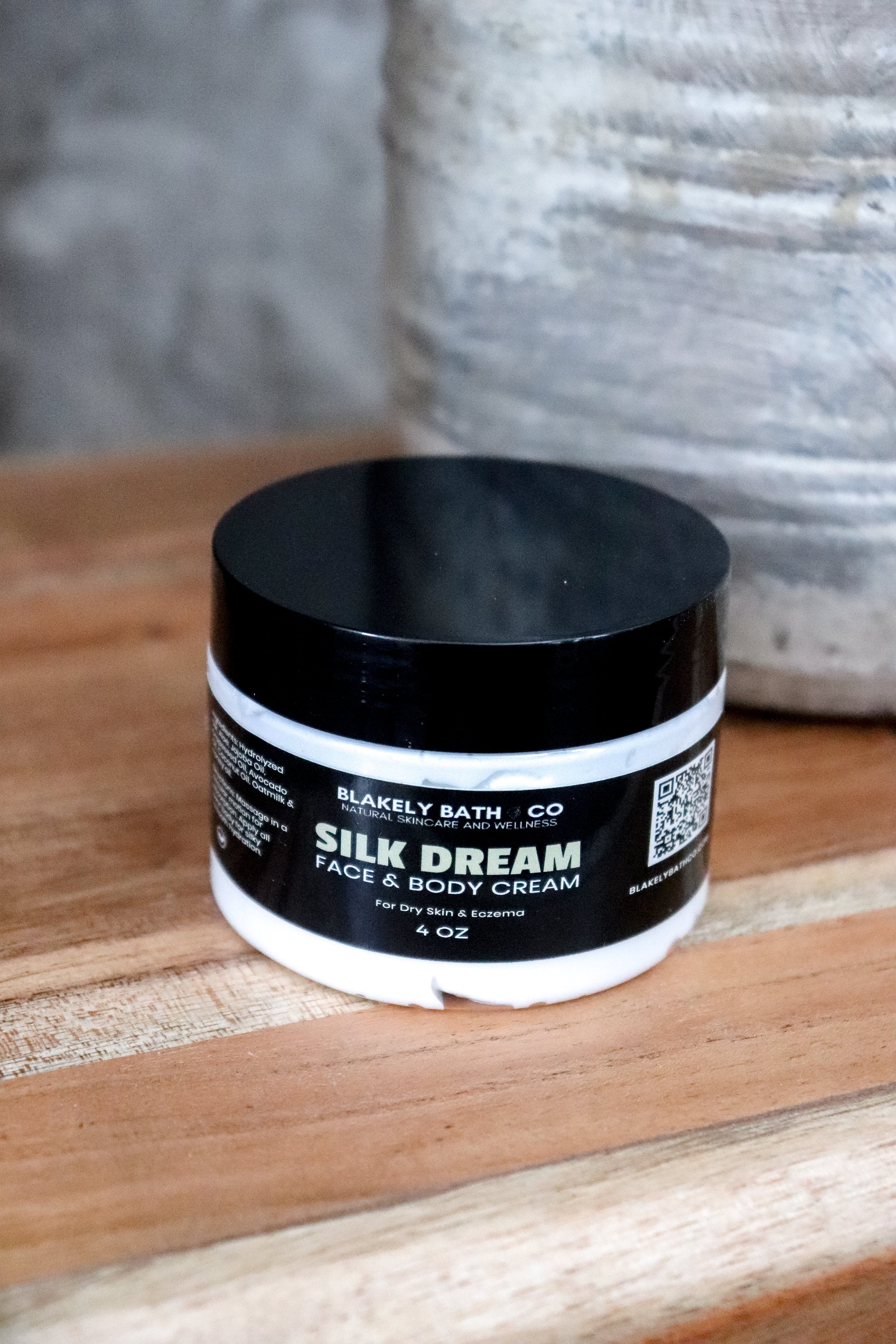 Jar of Blakely Bath Co Silk Dream Face & Body Cream on a wooden surface with a textured gray background best natural skincare moisturizer for radiant, hydrated, and silky smooth skin.